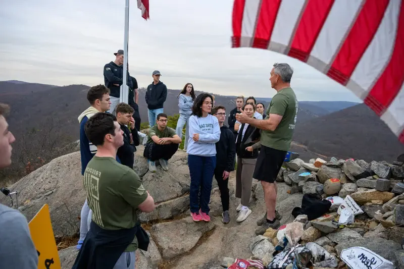 A group of students listens to a speaker atop a rocky summit. A corner of an American flag is visible in the foreground. The students wear a mix of casual clothing and Notre Dame apparel. The speaker, in an olive green shirt, gestures as he addresses them. A weathered flagpole stands behind the group with mountains visible in the background. A small rock cairn sits in the foreground left, adorned with painted stones and small signs.